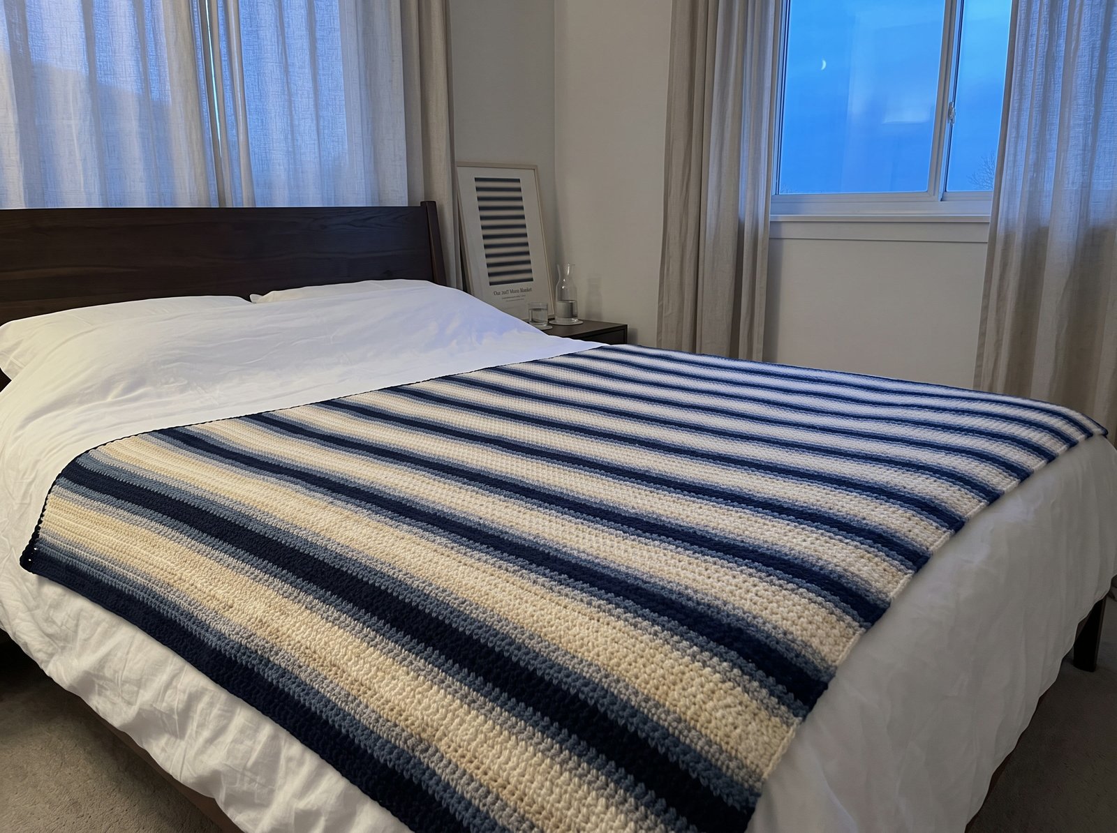 A crocheted moon phase blanket on a bed, stripes of navy, blue-grey and cream rising and falling with the moon, blue dusk at the window